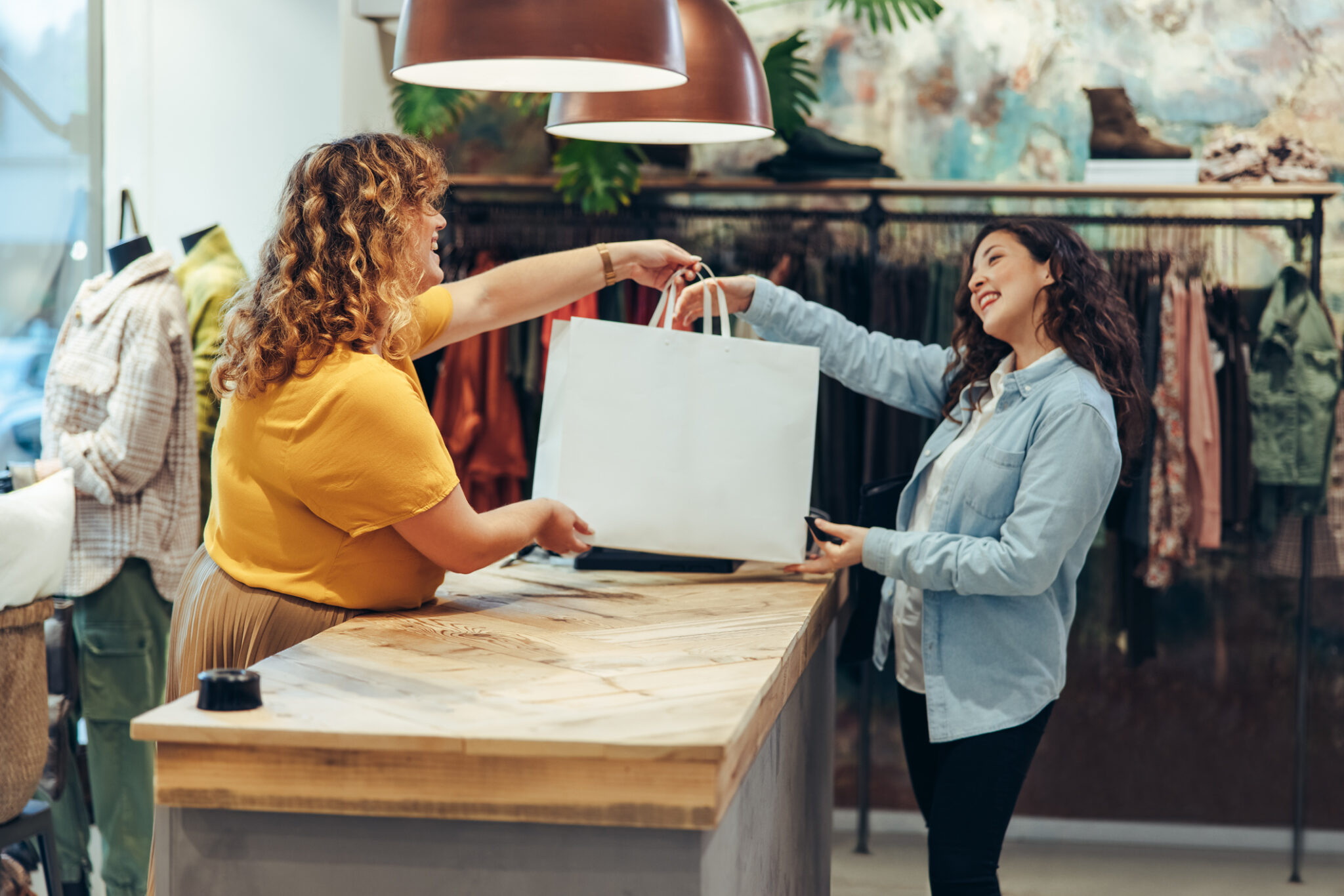 Friendly seller of clothing store giving shopping bags to satisfied female customer. Fashion store owner handing over the shopping bag to a female customer at checkout counter.